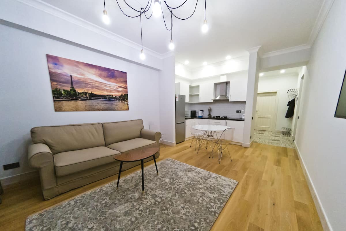 Una sala de estar con sofá, una mesa de centro y alfombra. Hay un cuadro en la pared y una cocina al fondo.