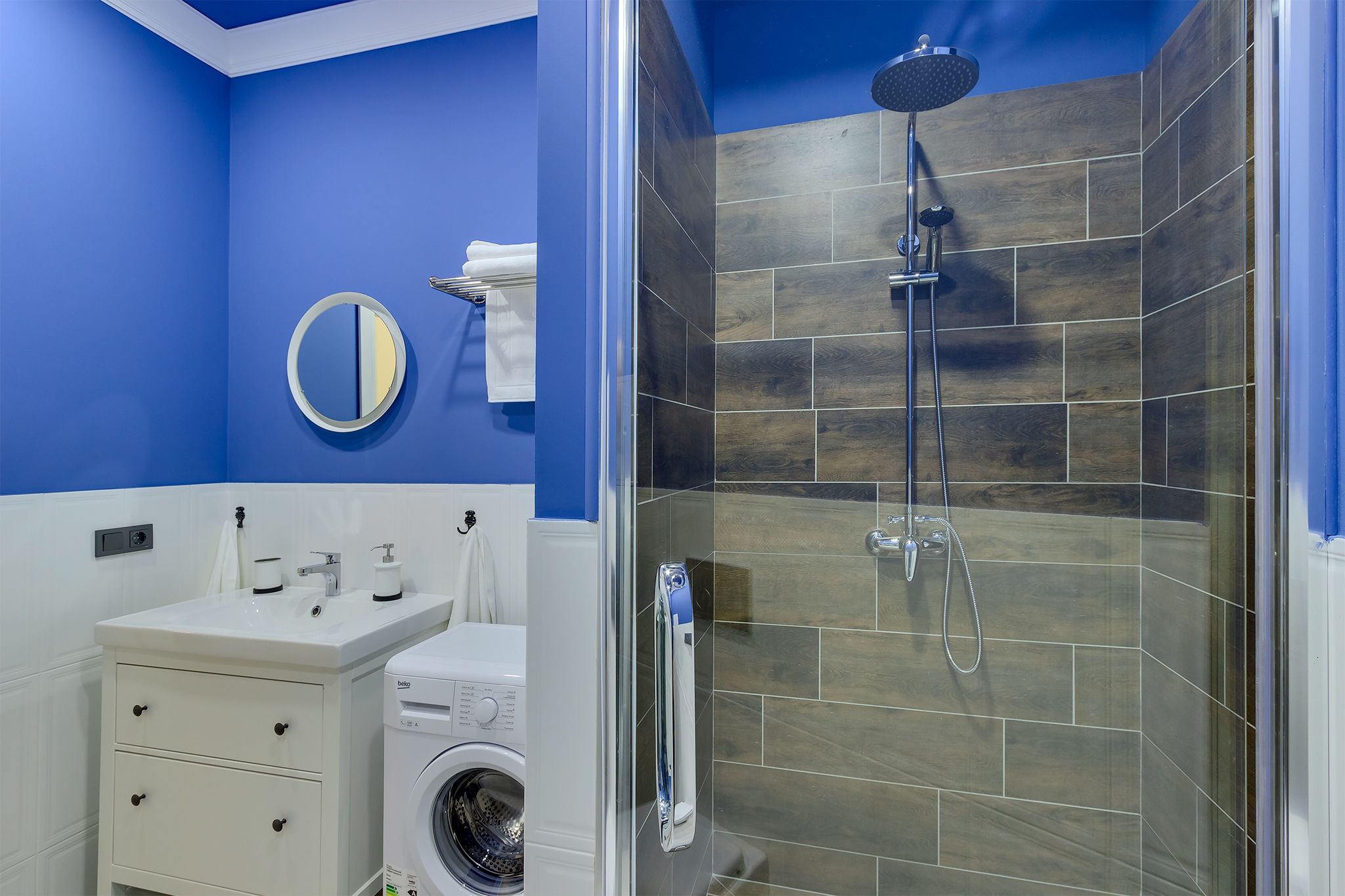 A bathroom with a blue wall, a round mirror, a white sink, a washing machine, and a shower with wooden tiles.