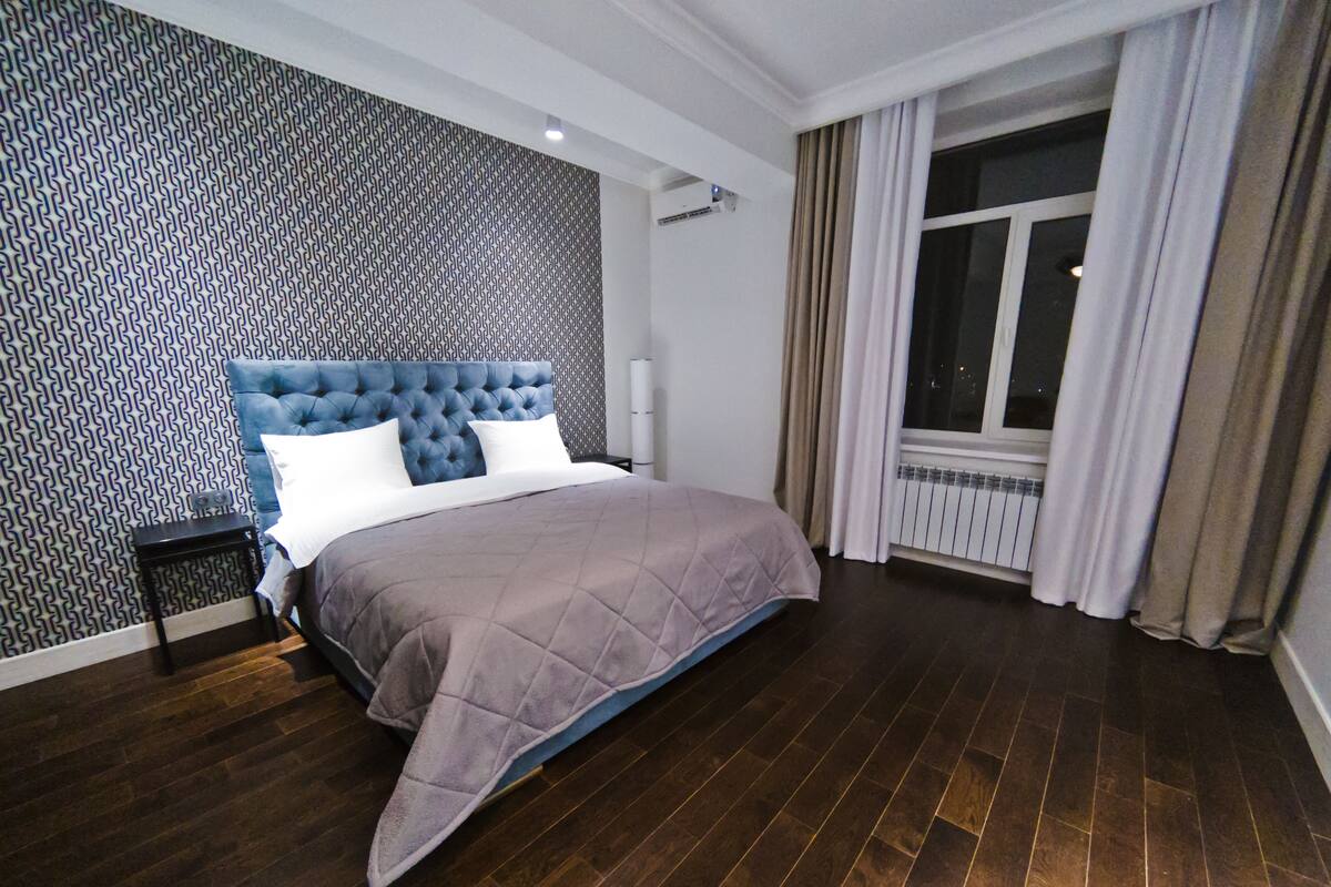 Un dormitorio con cabecera capitonada, una cama grande con colcha acolchada, pared con diseño, ventana con cortinas y un radiador.