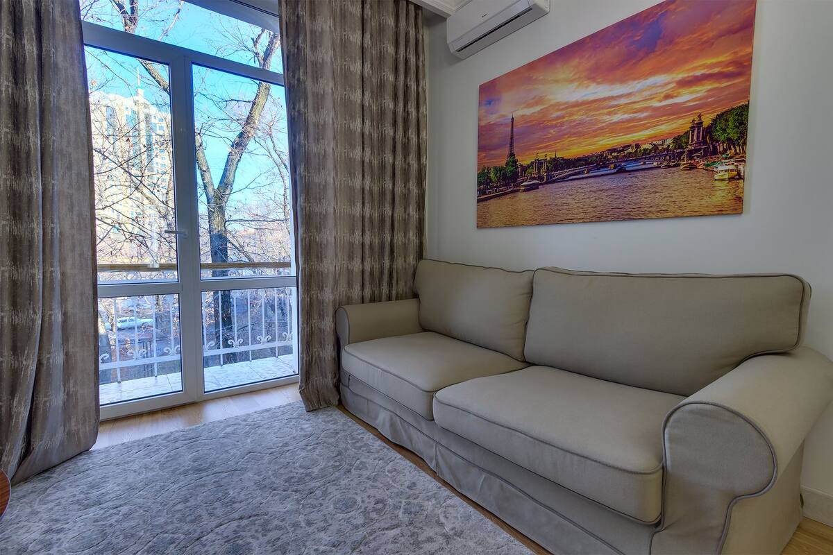 Una habitación con un sofá color beige, un ventanal con cortinas, un cuadro de un atardecer y un radiador.