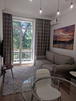 A living room with a sofa, a glass table, and a chair. There is a painting on the wall and a TV mounted on the wall.