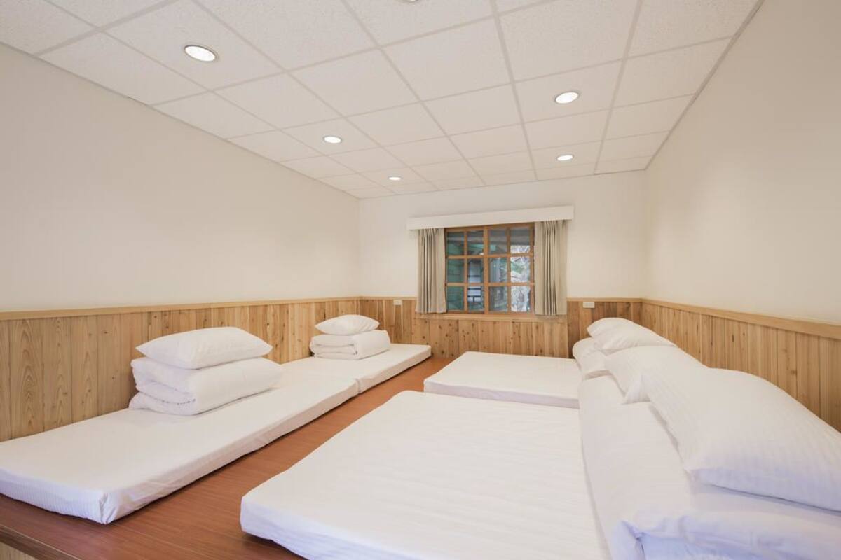 A room with three single beds arranged in a row, wooden paneling on the walls, a window with curtains, and recessed ceiling lighting.