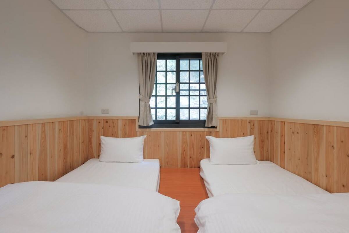 A room with two beds, wooden paneling, a window with curtains, and a view of trees outside.