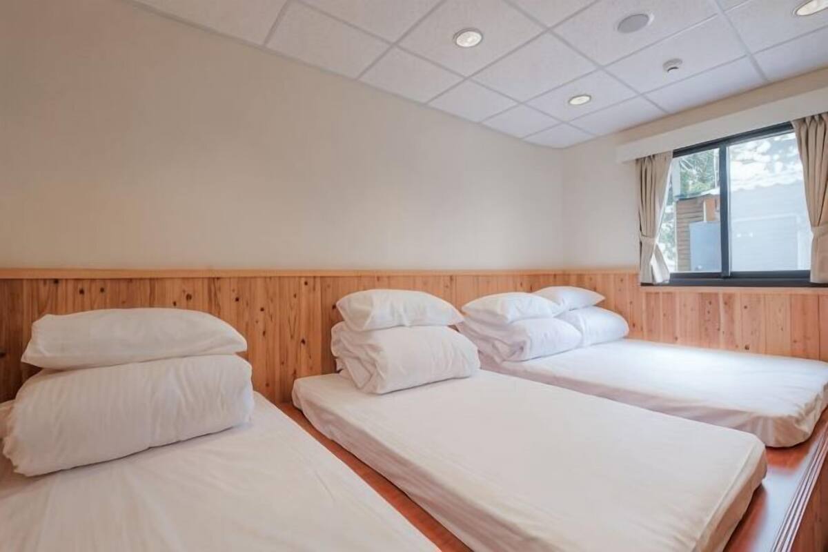 A room with two beds, wooden headboard, white bedding, a window with curtains, and a ceiling with recessed lighting.