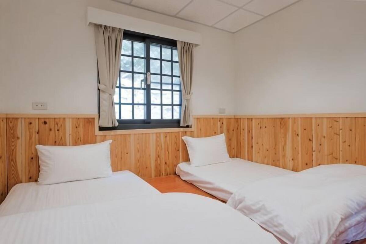 A room with two beds, wooden paneling, a window with curtains, and a white ceiling.