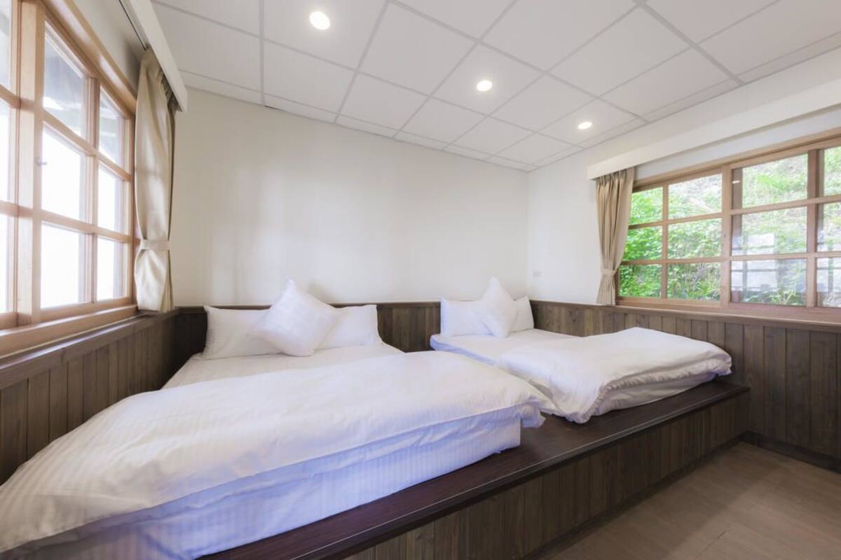 A hotel room with two beds, wooden paneling, large windows, and a view of greenery outside.