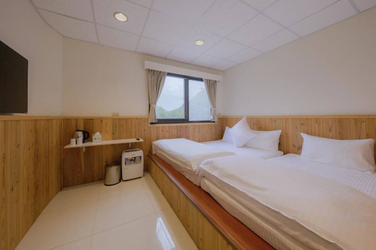 A hotel room with two single beds, a wooden headboard, a window with curtains, a shelf with toiletries, and a tiled floor.