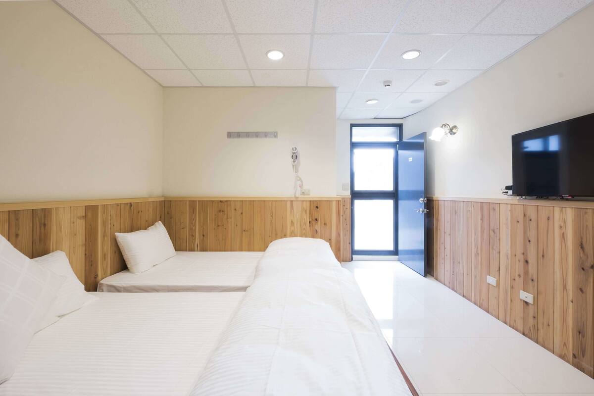 A hotel room with two beds, wooden paneling, a television, and a door leading to a bright area.