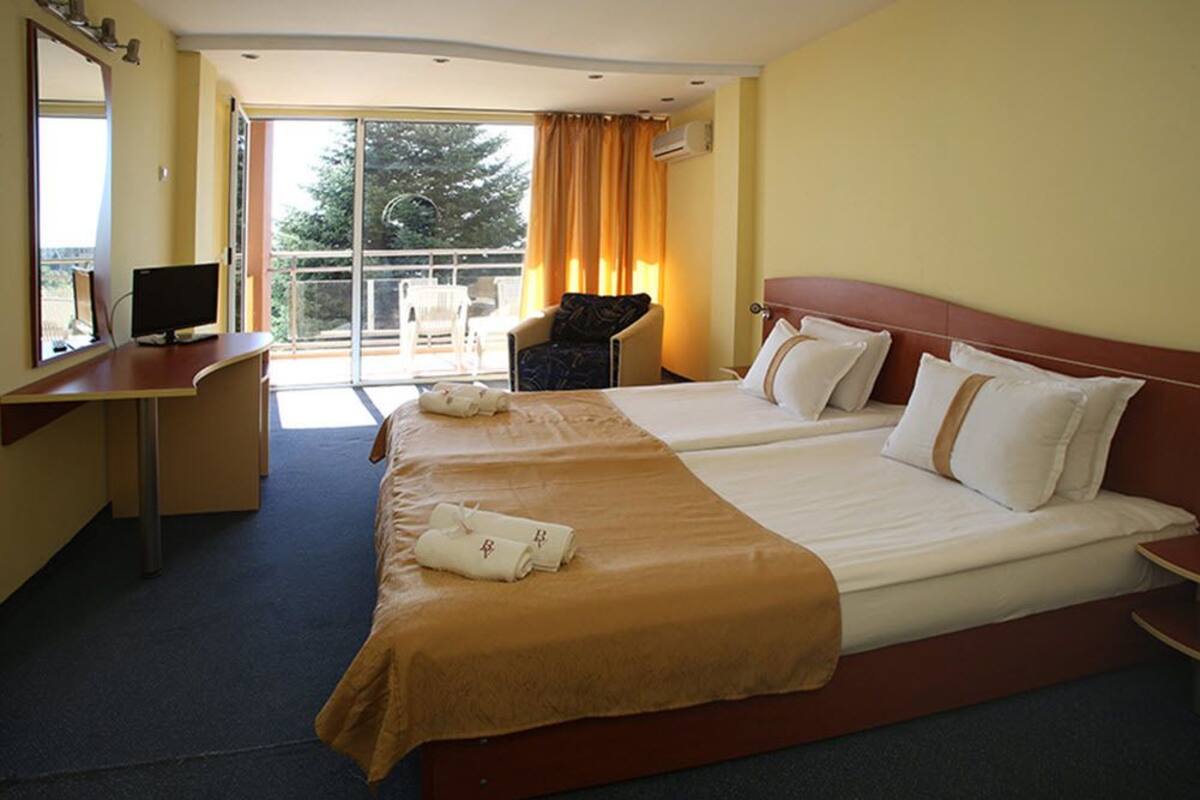 A hotel room with two beds, a desk with a computer, a chair, and a balcony with a view.