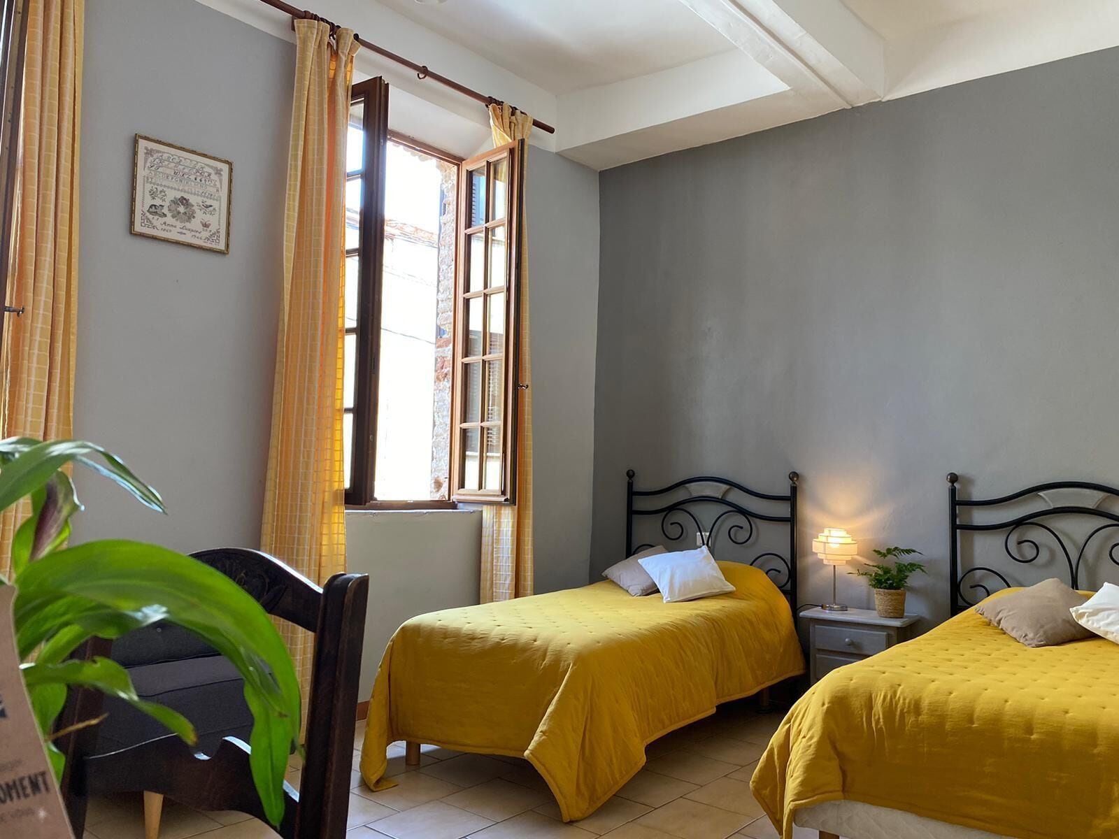 A room with two beds, a window with curtains, a bedside table with a lamp, and a framed picture on the wall.