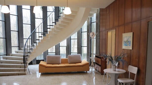 A spacious lobby with a curved staircase, a modern sofa with cushions, a small round table with a chair, and a potted plant. - Tango (Golden Sands)