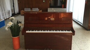 A wooden piano in a lobby with a reception desk, chairs, and a potted plant. - Tango (Golden Sands)