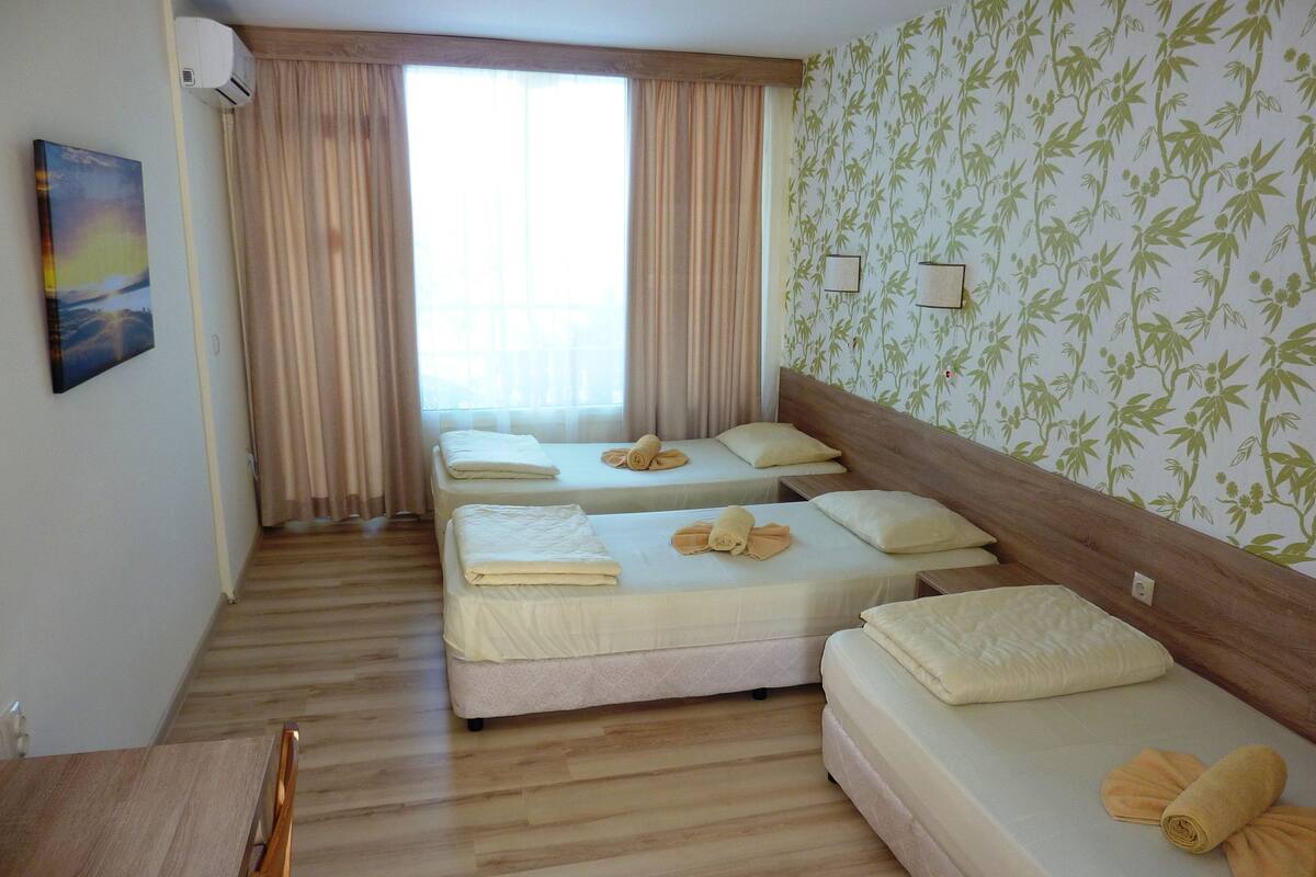 A hotel room with three beds, a wooden floor, and a view of the outdoors through large windows.
