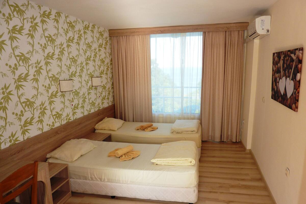 A hotel room with two beds, a wooden desk, a window with curtains, and an air conditioning unit on the wall.