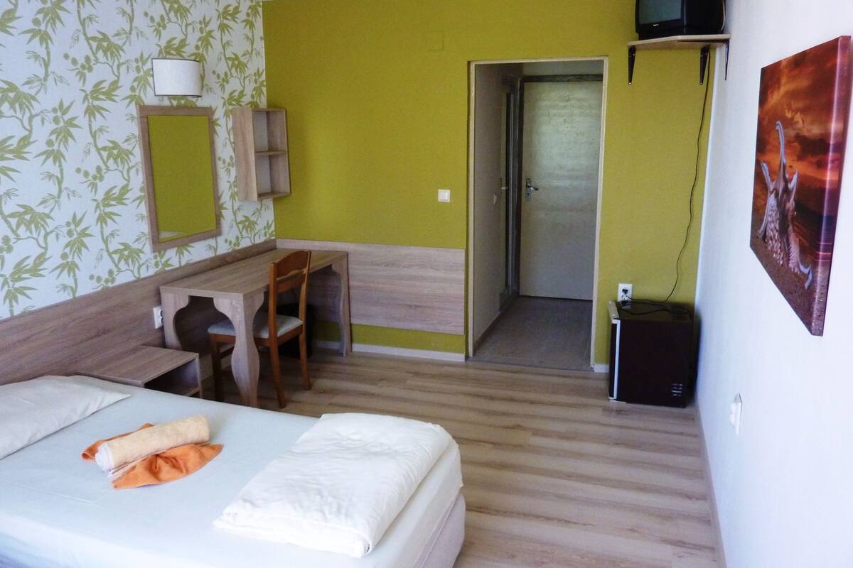 A hotel room with a bed, a desk, a chair, a TV, and a door leading to a bathroom.