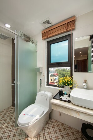 Et moderne badeværelse med brusekabine i glas, toilet med skilt med teksten "Rengjort og desinficeret“, et vindue med udsigt til bygninger og træer, og en vask med spejl og en vase med gule blomster.