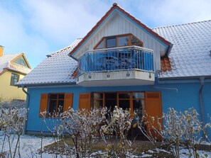 Kaksikerroksinen talo, jossa on parveke, lumipeitteinen katto ja puutarha, jossa on paljaita puita.