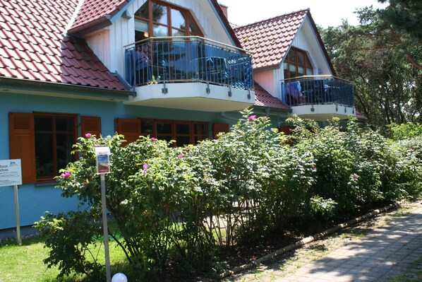 A two-story house with a balcony, a garden with pink flowers, and a sign on the left.