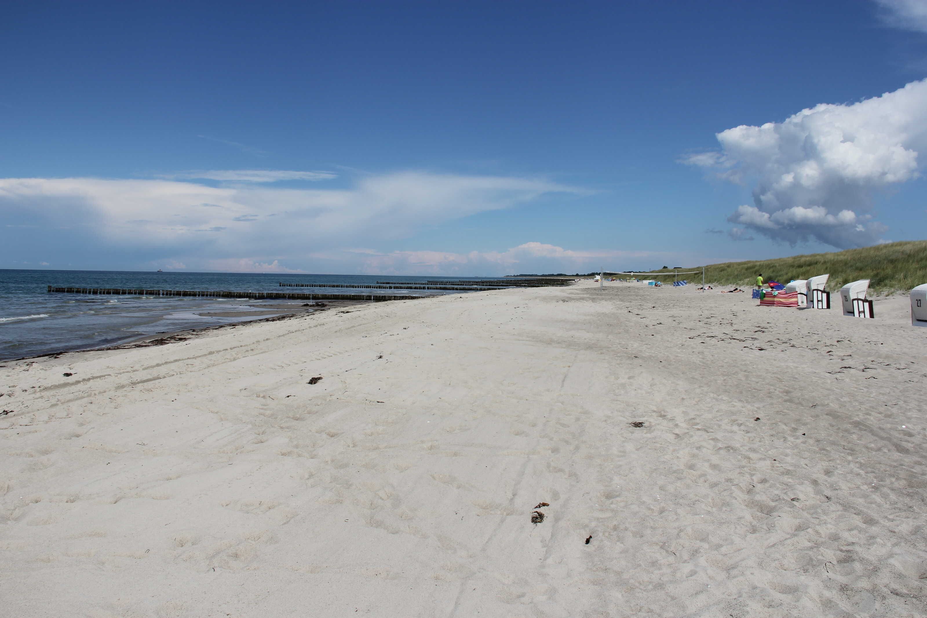 A beach with a wooden pier extending into the water, white beach chairs, and a dune in the background.