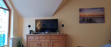 A living room with a TV, wooden cabinets, a sofa, a table with a bottle and glasses, and a painting on the wall.