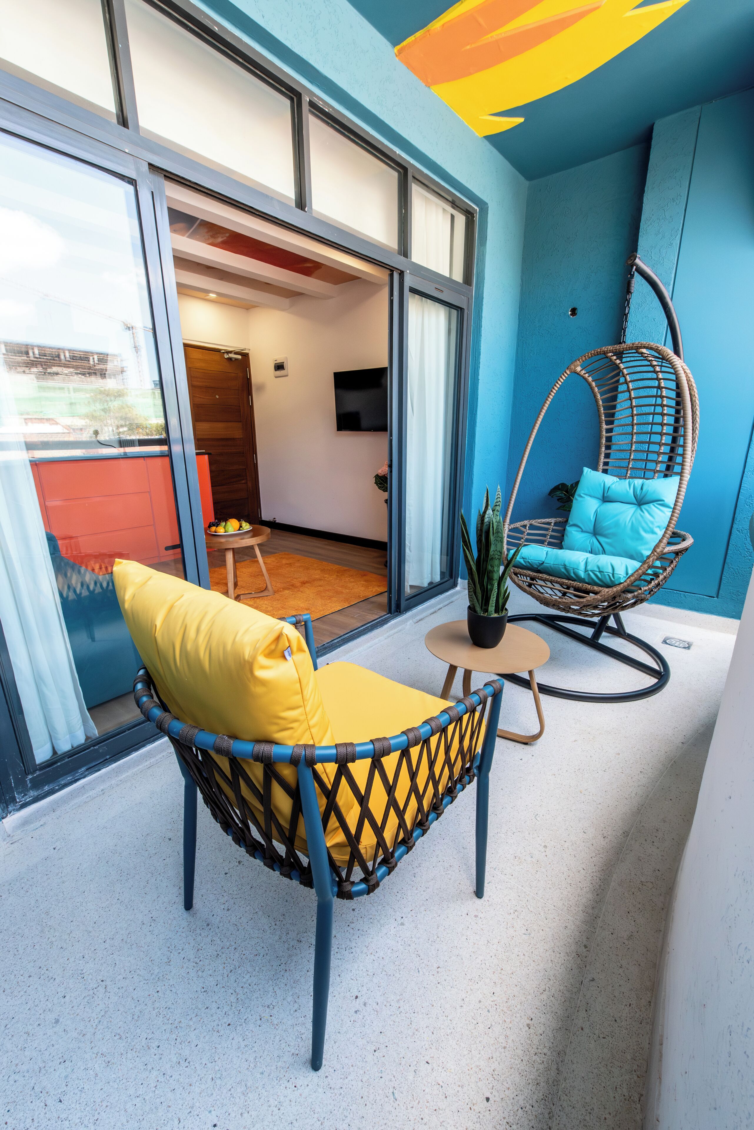 A balcony with a wicker chair and a yellow sofa, a small table with a potted plant, and a view of the interior space.