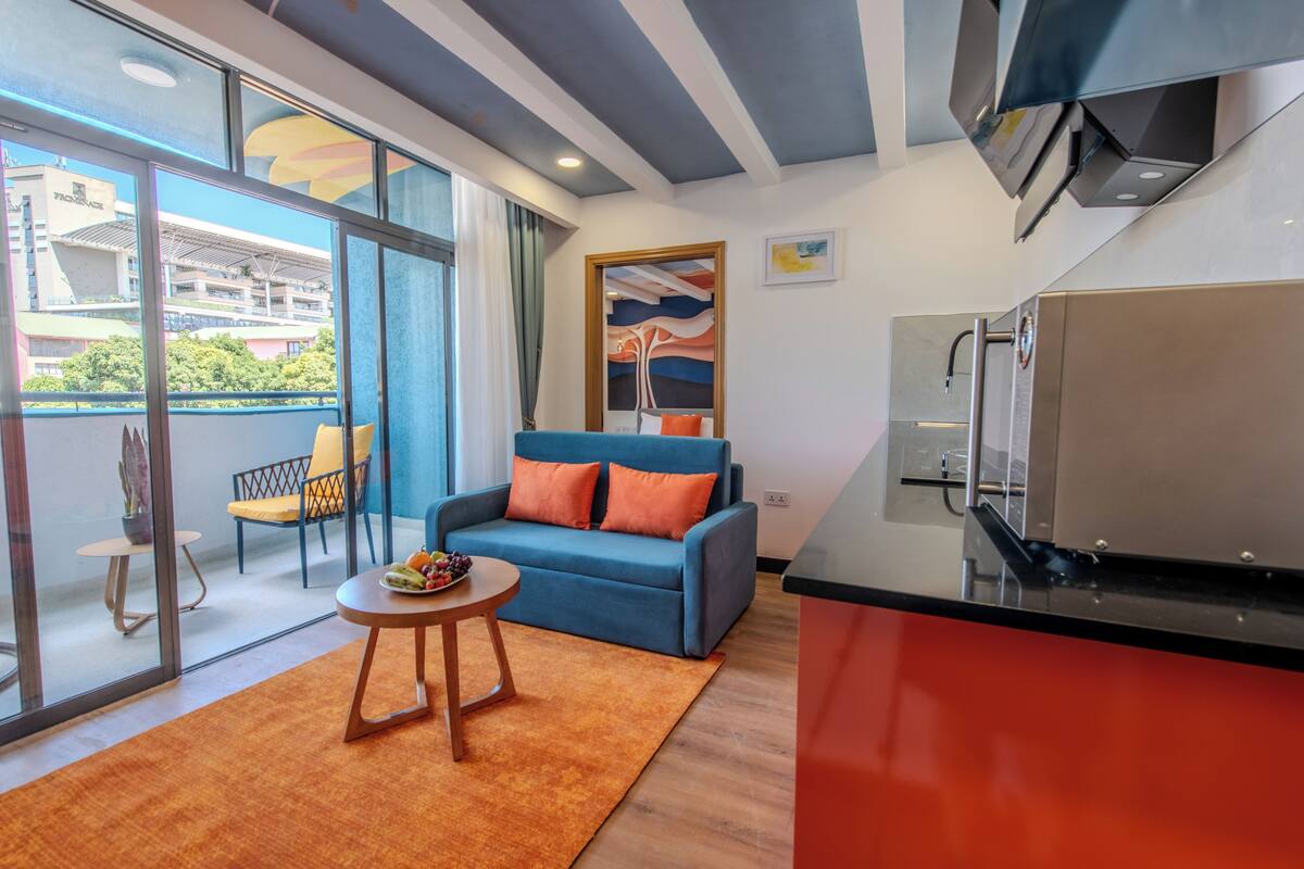 A modern living room with a blue sofa, orange cushions, a small wooden table with fruit, and a view of a building through large windows.