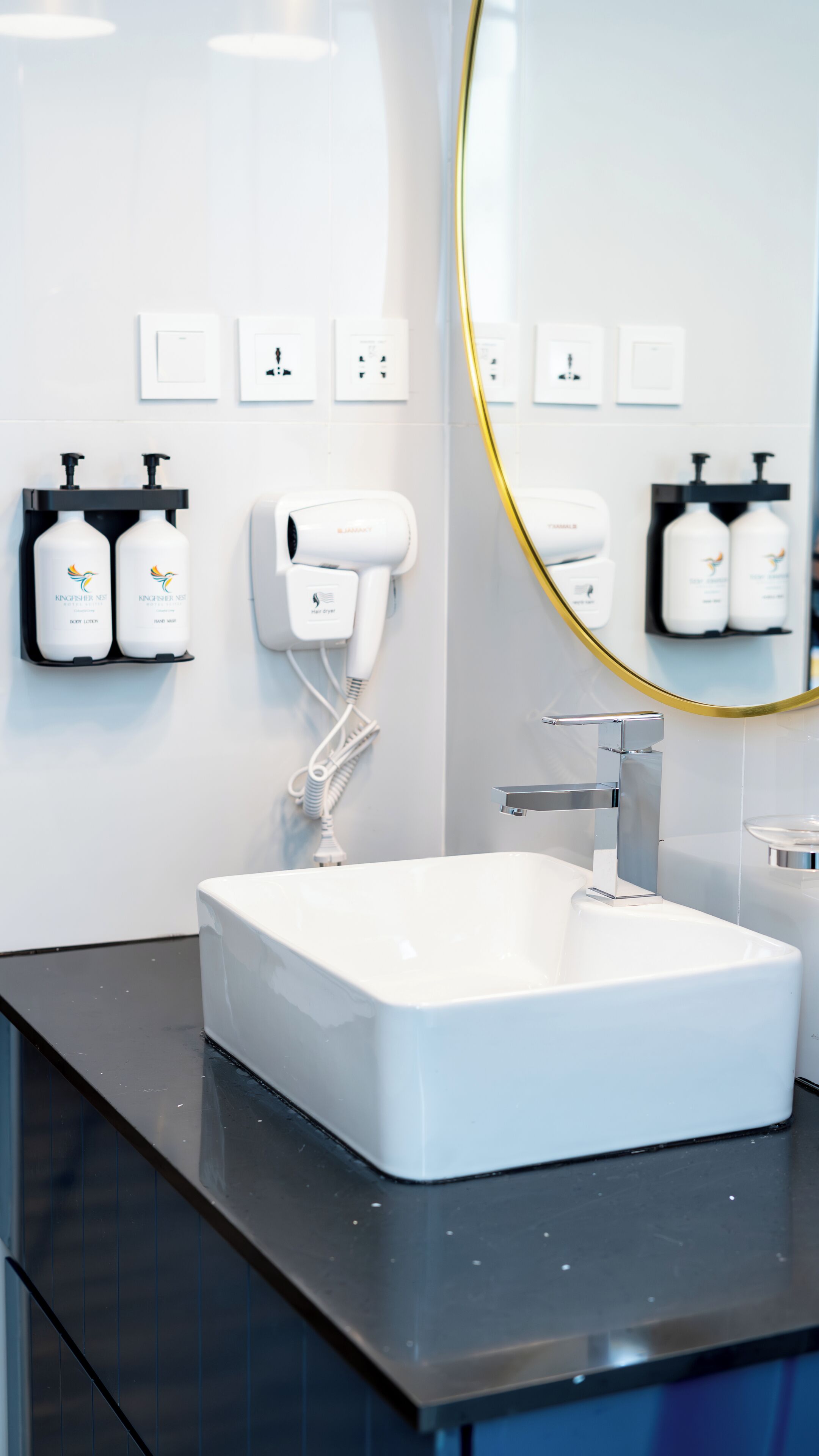 Een moderne badkamer met een wit wastafel, een haardroger en handzeepdispensers aan de muur.
