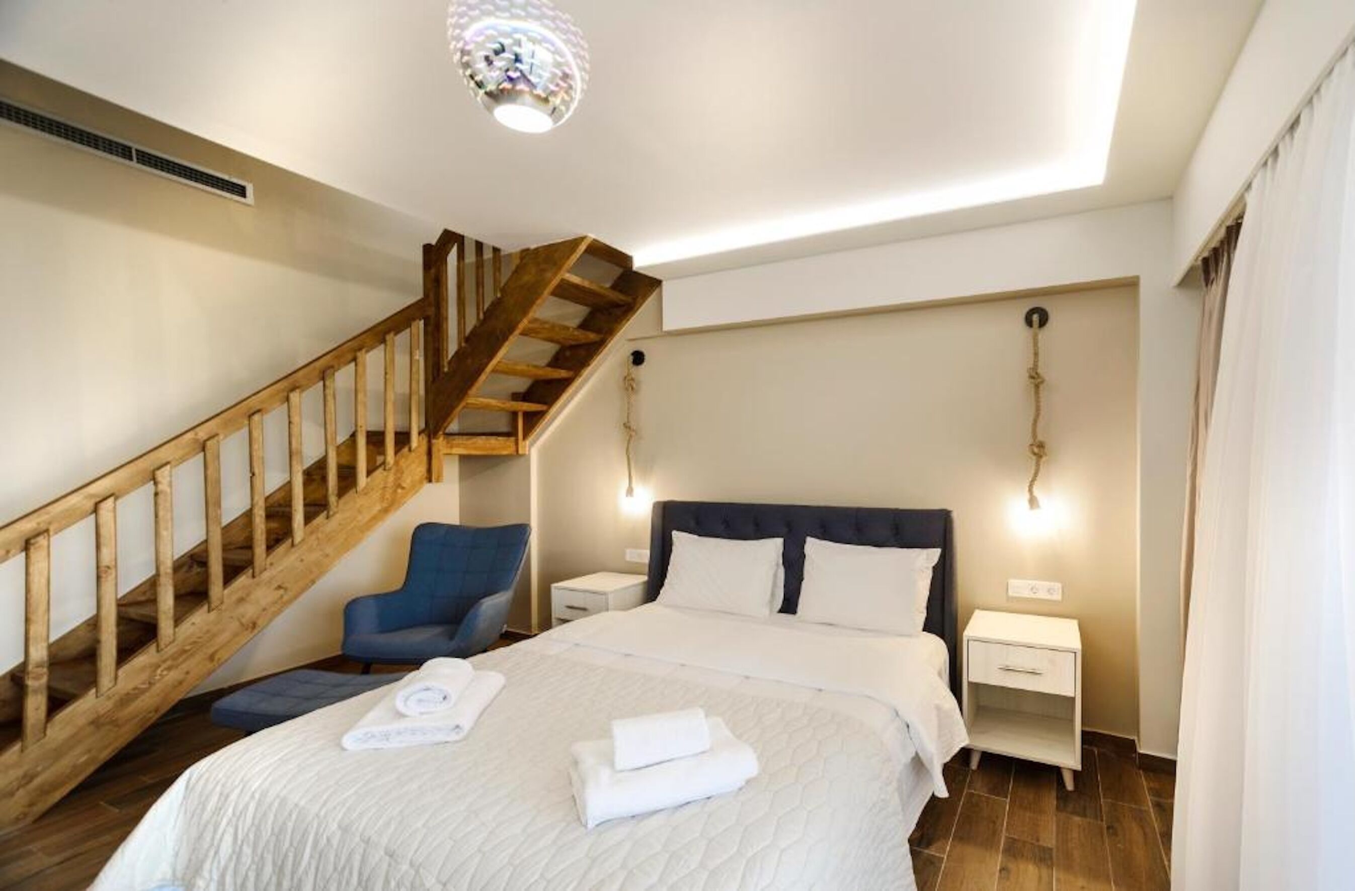 A bedroom with a wooden staircase, a bed with white linens, a blue chair, and bedside tables with lamps.