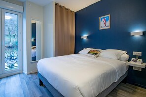A hotel room with a large bed, a window with a view of trees, a wall-mounted light, and a poster on the wall. - Hôtel Le Regis (Mont-Dore)