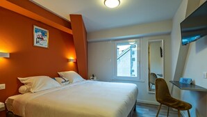 A hotel room with a bed, a chair, a desk, a TV, and a window with a view of buildings. - Hôtel Le Regis (Mont-Dore)