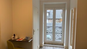 A narrow hallway with a door leading to a balcony with a metal railing. A small table with a chair and some items is placed against the wall. - Hôtel Le Regis (Mont-Dore)