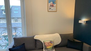 A living room with a sofa, a poster on the wall, and a view of the outside. - Hôtel Le Regis (Mont-Dore)