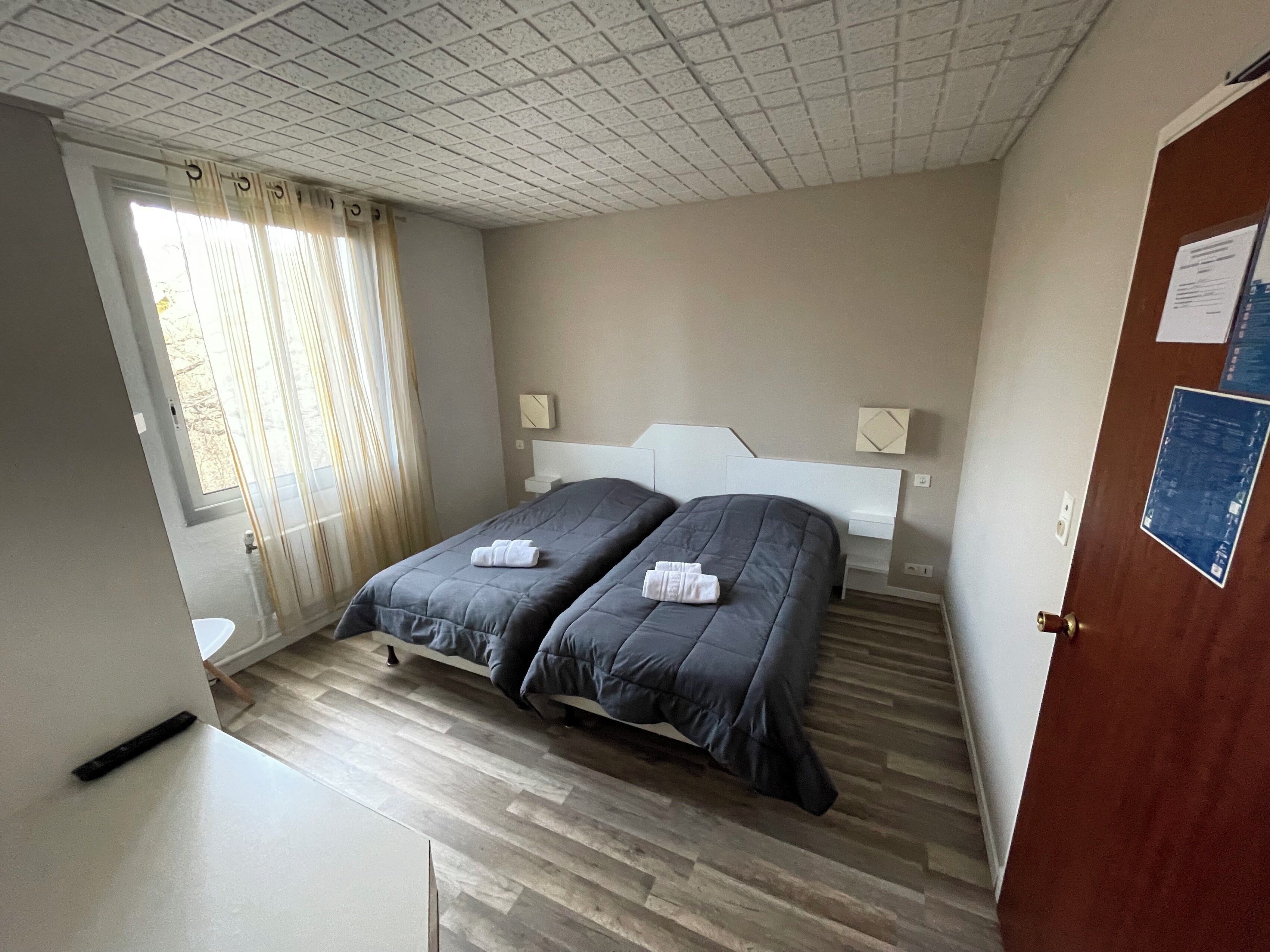 A bedroom with two beds, a wooden floor, a window with curtains, and a door with a notice.