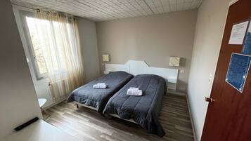 A bedroom with two beds, a wooden floor, a window with curtains, and a door with a notice.