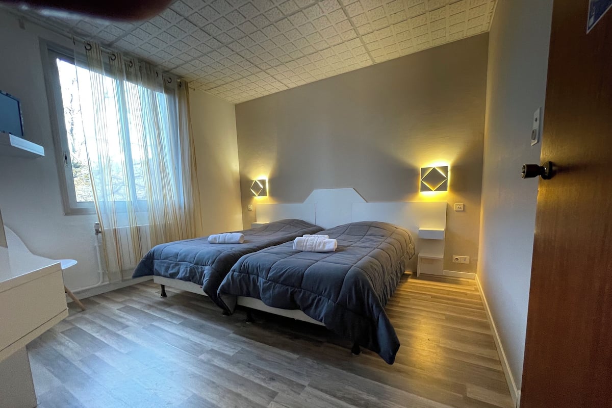 Un dormitorio con una cama doble, dos mesitas de noche con lámparas, un ventanal con cortinas y suelo de madera.