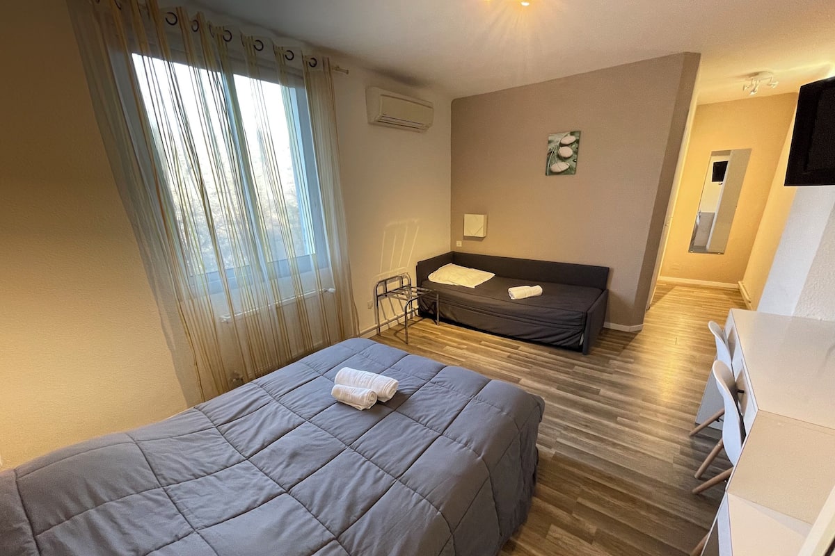 Habitación de hotel con cama, escritorio, silla, ventana con cortinas, aire acondicionado de pared y una obra de arte pequeña.
