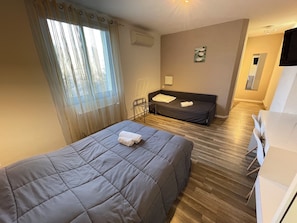 A hotel room with a bed, a desk, a chair, a window with curtains, a wall-mounted air conditioner, and a small artwork.