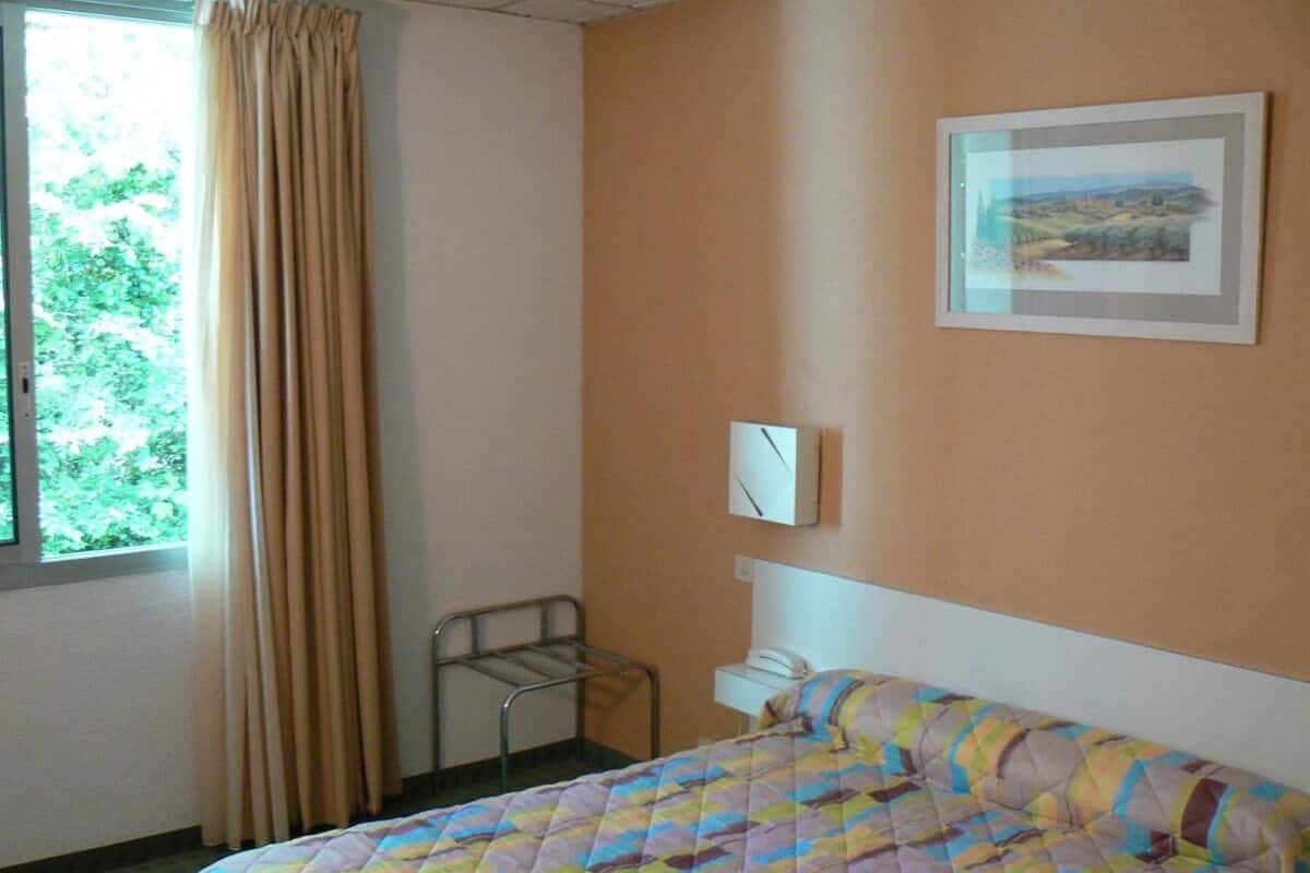 Habitación de hotel con cama, mesita de noche, una silla, ventana con cortinas y un cuadro en la pared.