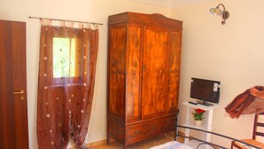 A bedroom with a wooden wardrobe, a television on a stand, a bed with a floral bedspread, and a chair. - Casale Racalmare (Grotte)