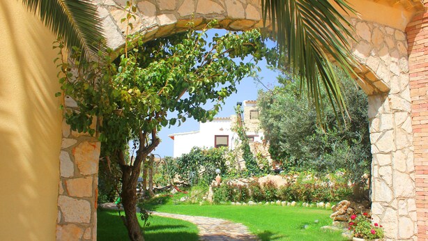 A stone archway leading to a garden with a pathway, greenery, and a building in the background. - Casale Racalmare (Grotte)