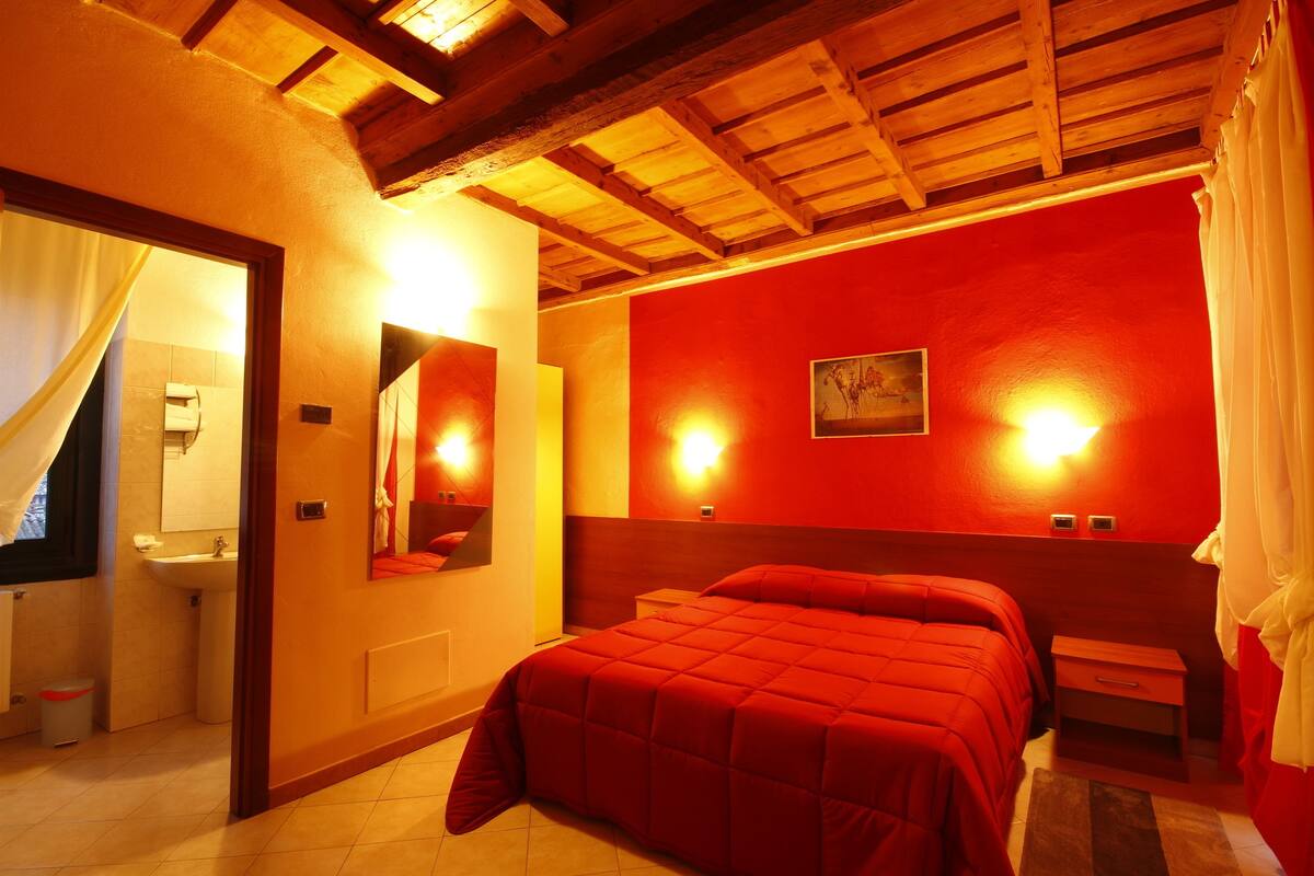 Une chambre à coucher avec une couvre-lit rouge, des poutres apparentes en bois au plafond, une table de chevet et un luminaire fixé au mur.