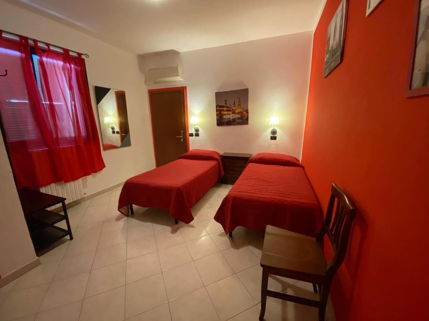 A hotel room with two single beds, a chair, a small table, a door, an air conditioning unit, and a wall-mounted lamp.