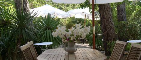 A round wooden table with a white umbrella, surrounded by striped chairs, in a lush garden setting.