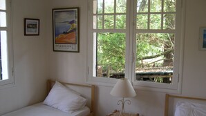 Una habitación pequeña y sencilla con dos camas individuales, una mesita con una lámpara y una ventana que da a zonas verdes.