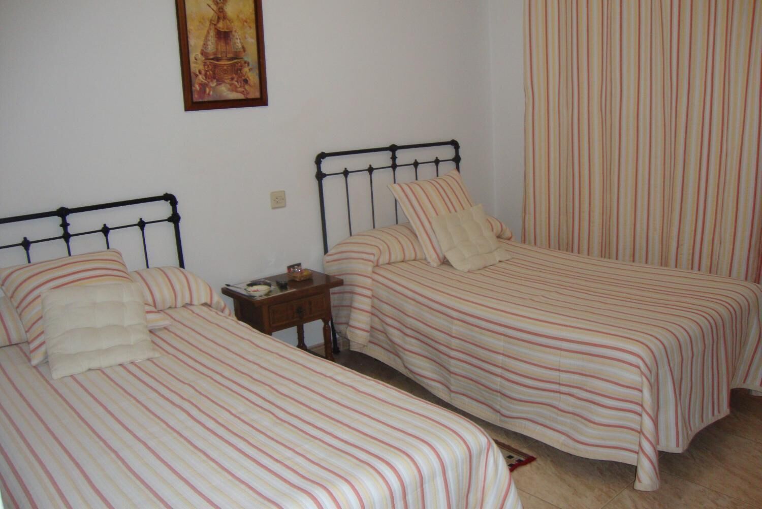 A room with two single beds, each with striped bedding and pillows. There is a small wooden nightstand with a lamp and a book. A painting hangs on the wall above the beds.