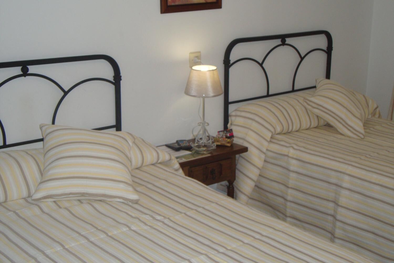 Habitación con dos camas, cada una con ropa de cama a rayas y cabeceros de metal negro. Entre las camas, un mesita de noche con una lámpara y un pequeño objeto decorativo.