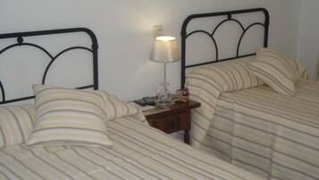 Quarto com duas camas, cada uma com roupa de cama listrada e cabeceiras de metal preto. Entre as camas, um criado-mudo com abajur e um pequeno objeto decorativo.