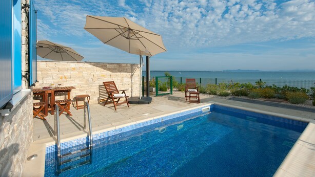 Una piscina con vistas al mar, rodeada de muros de piedra y mobiliario de madera.