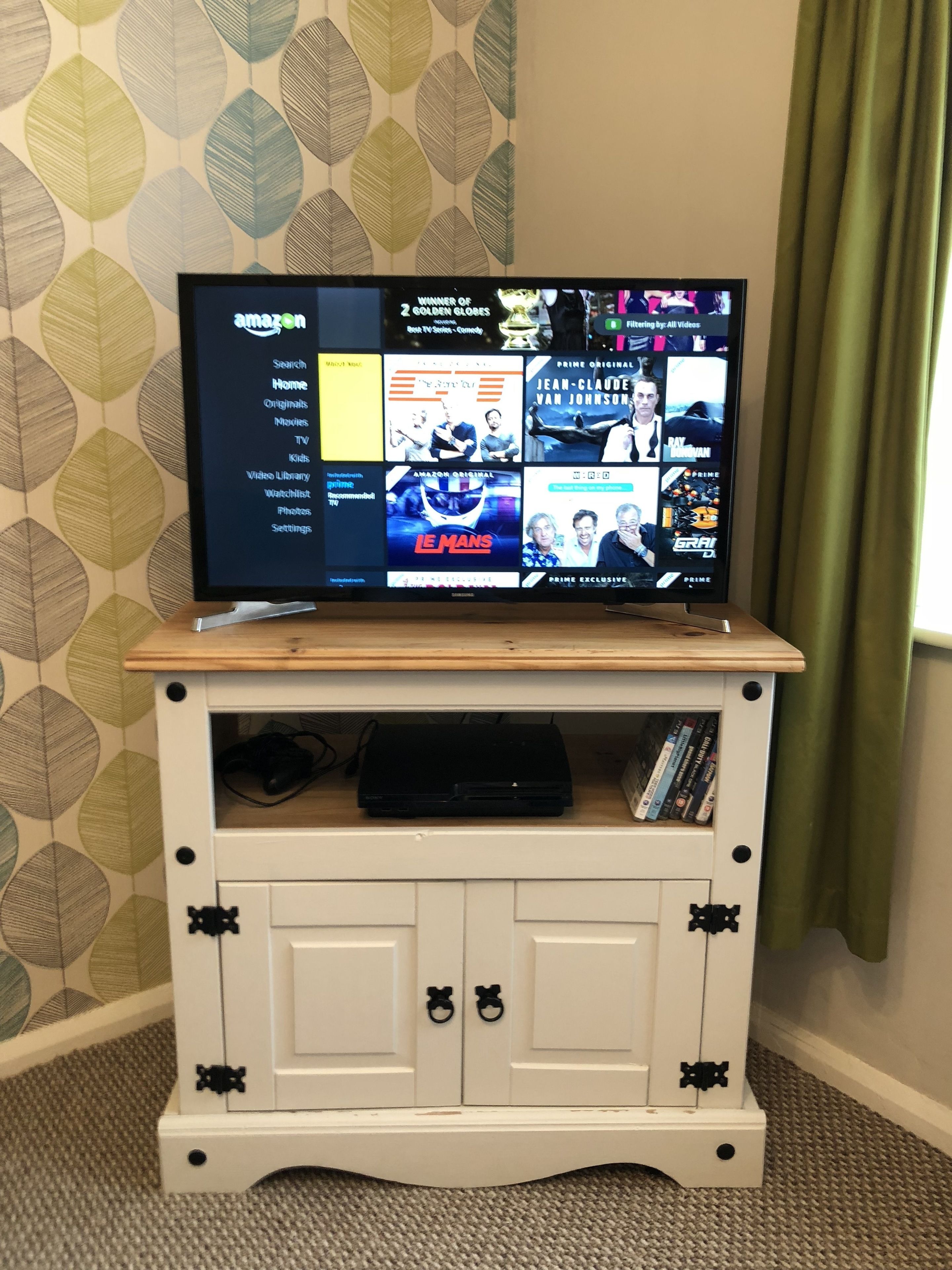 A flat-screen TV on a wooden TV stand with a leaf-patterned wallpaper in the background.