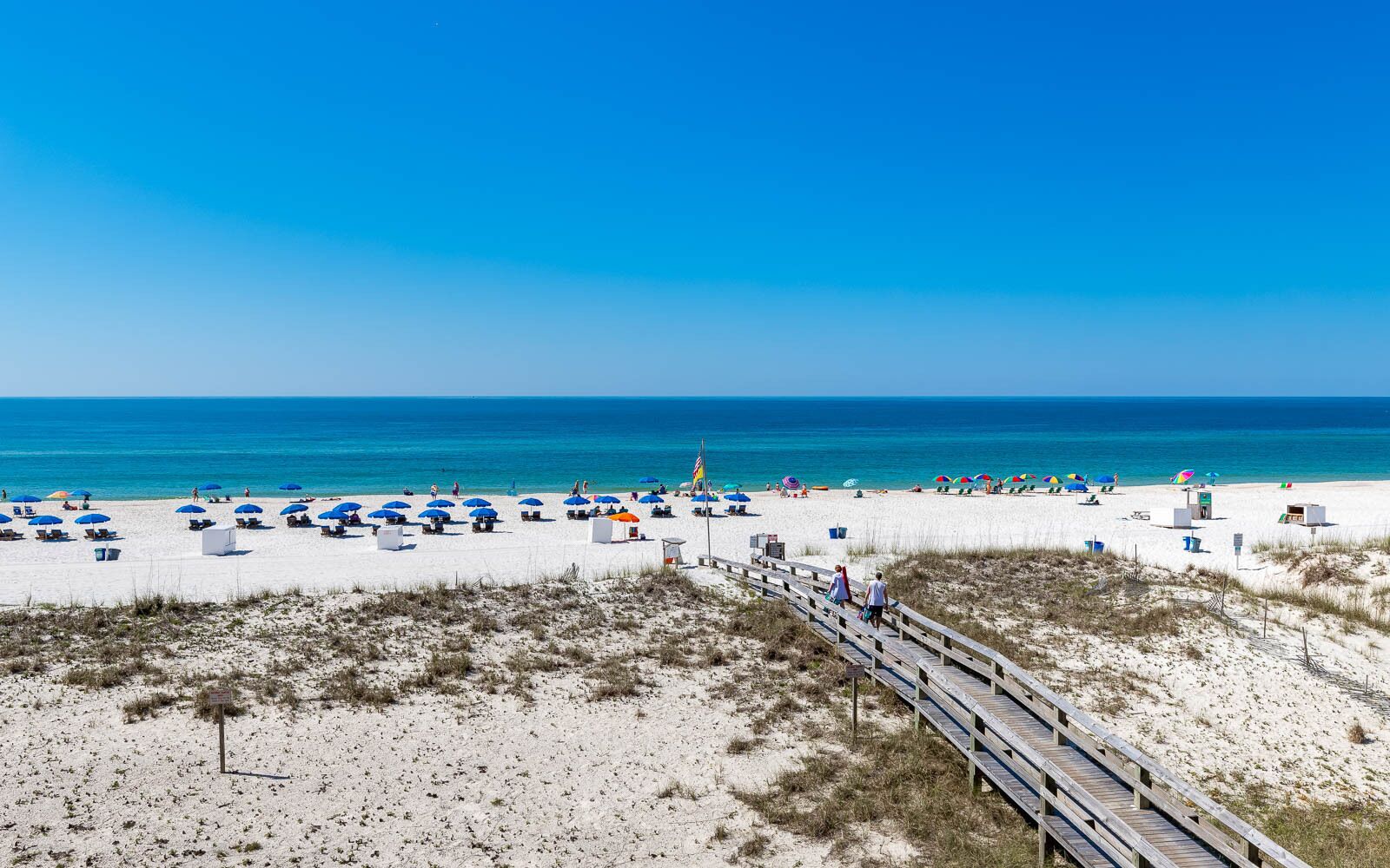 A beach with white sand, blue umbrellas, and a wooden boardwalk.
